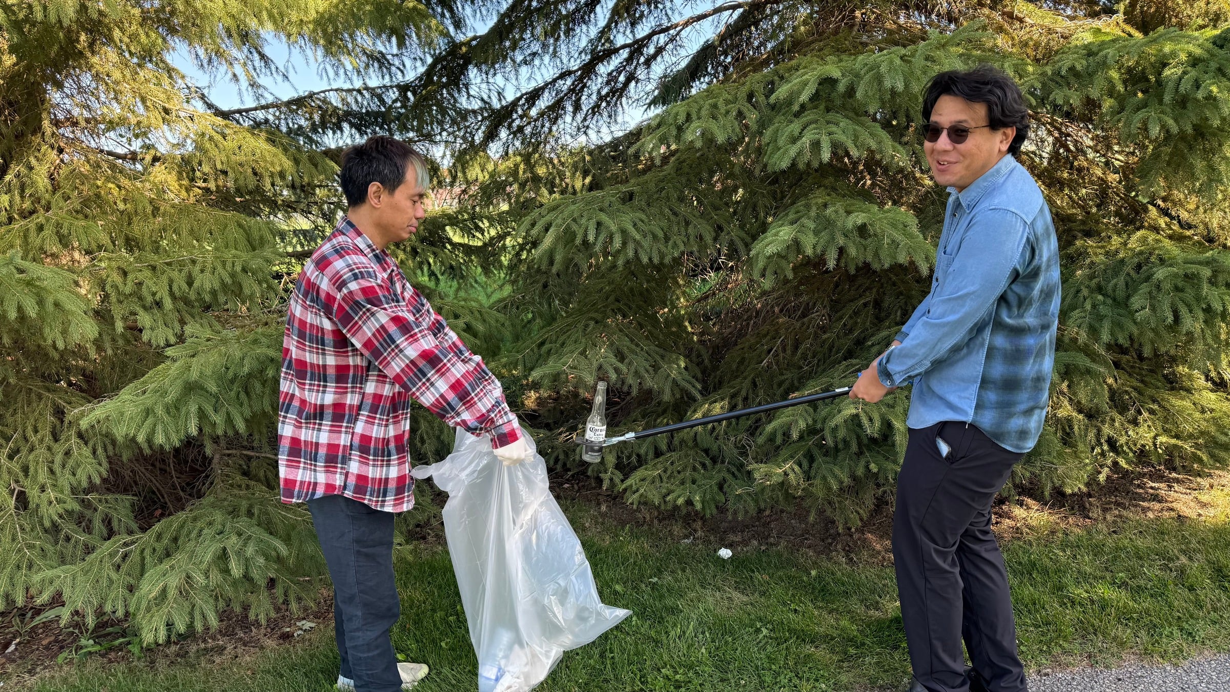 Two volunteers picking up a bottle with a litter grabber beside evergreen trees